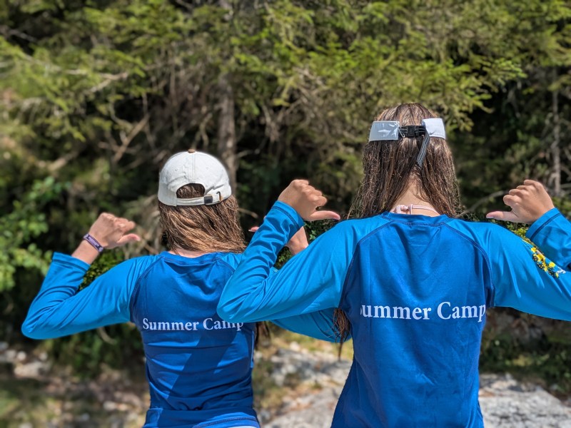 Two campers outdoors, pointing to the “Summer Camp” lettering on the backs of their blue shirts.